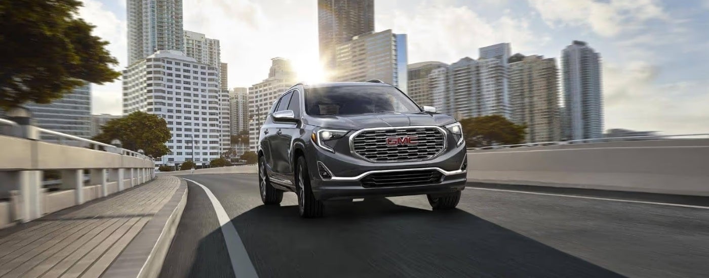 A silver 2018 GMC Terrain Denali is driving on a city street past buildings.